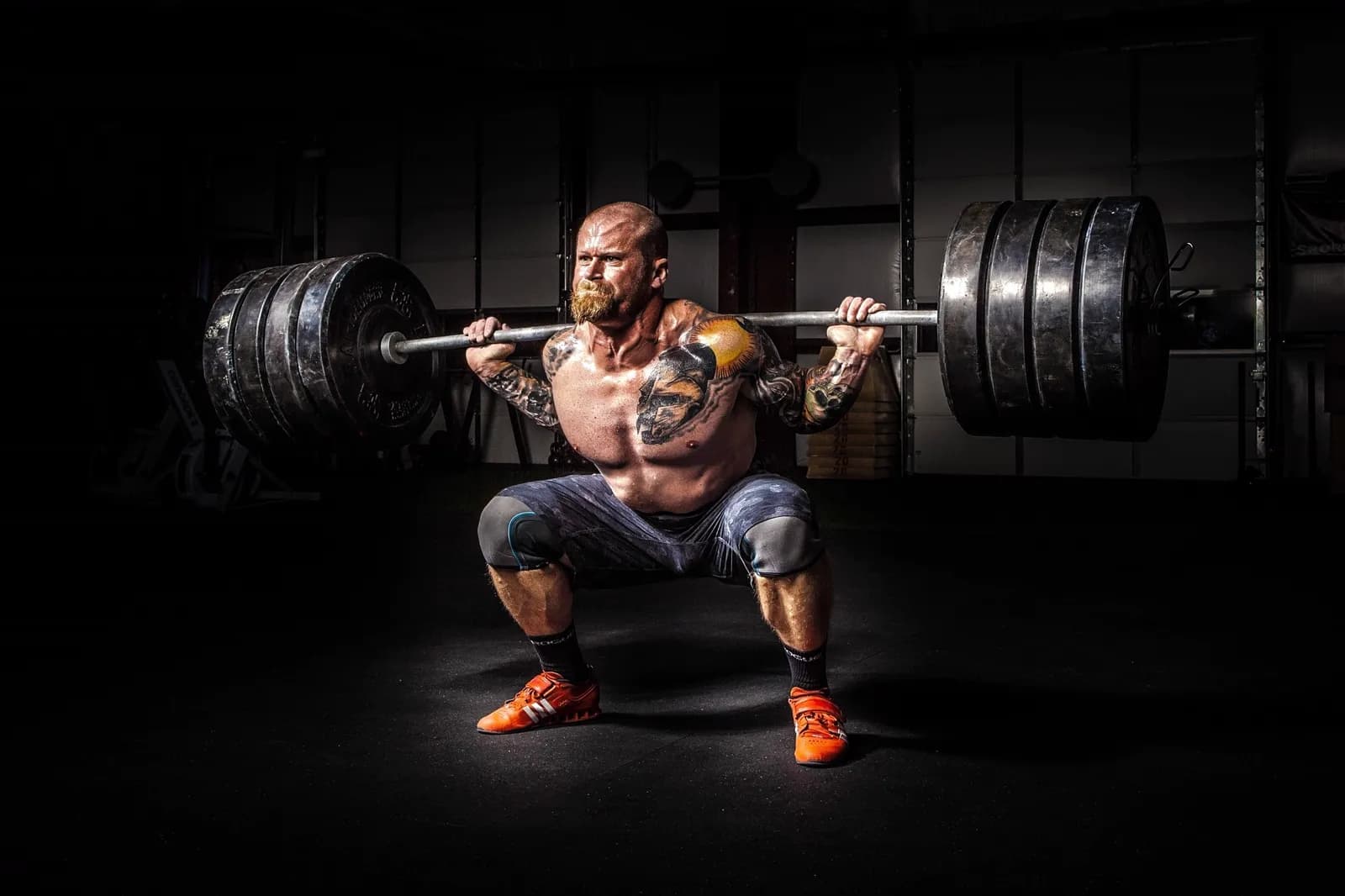 A lifter at the bottom of a heavy back squat under a barbell loaded with bumper plates, in a darkened gym.