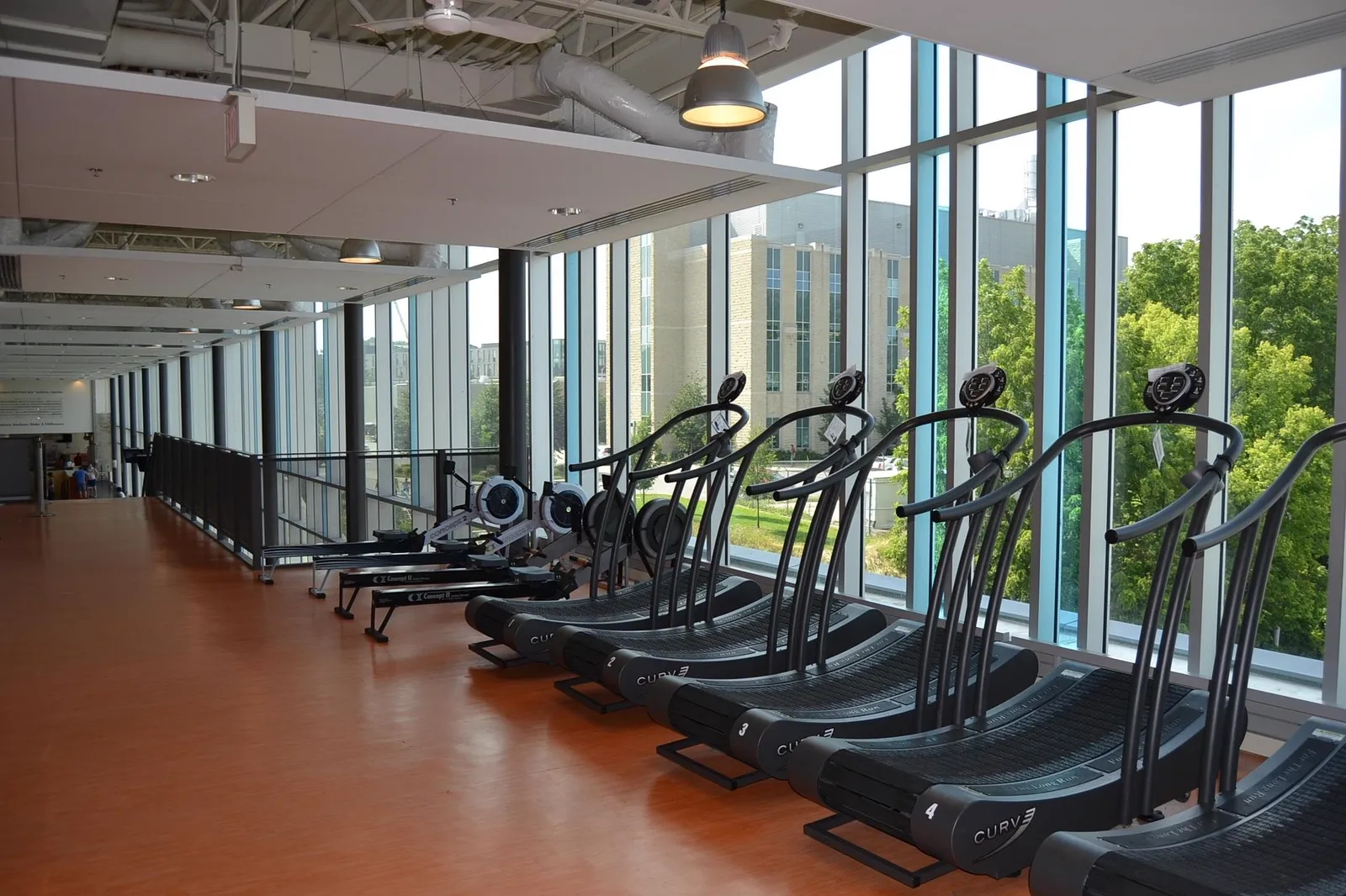 A row of curved manual treadmills and rowing machines along a wall of full-height windows on a cardio floor.