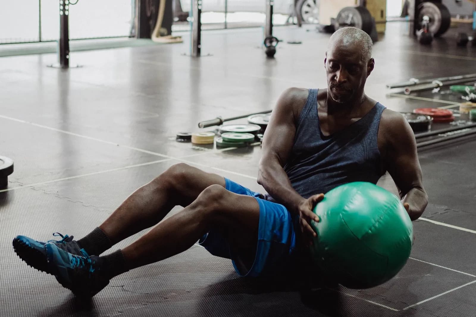 A person seated on a gym floor mid-twist holding a green medicine ball, with loose weight plates and a barbell behind them.