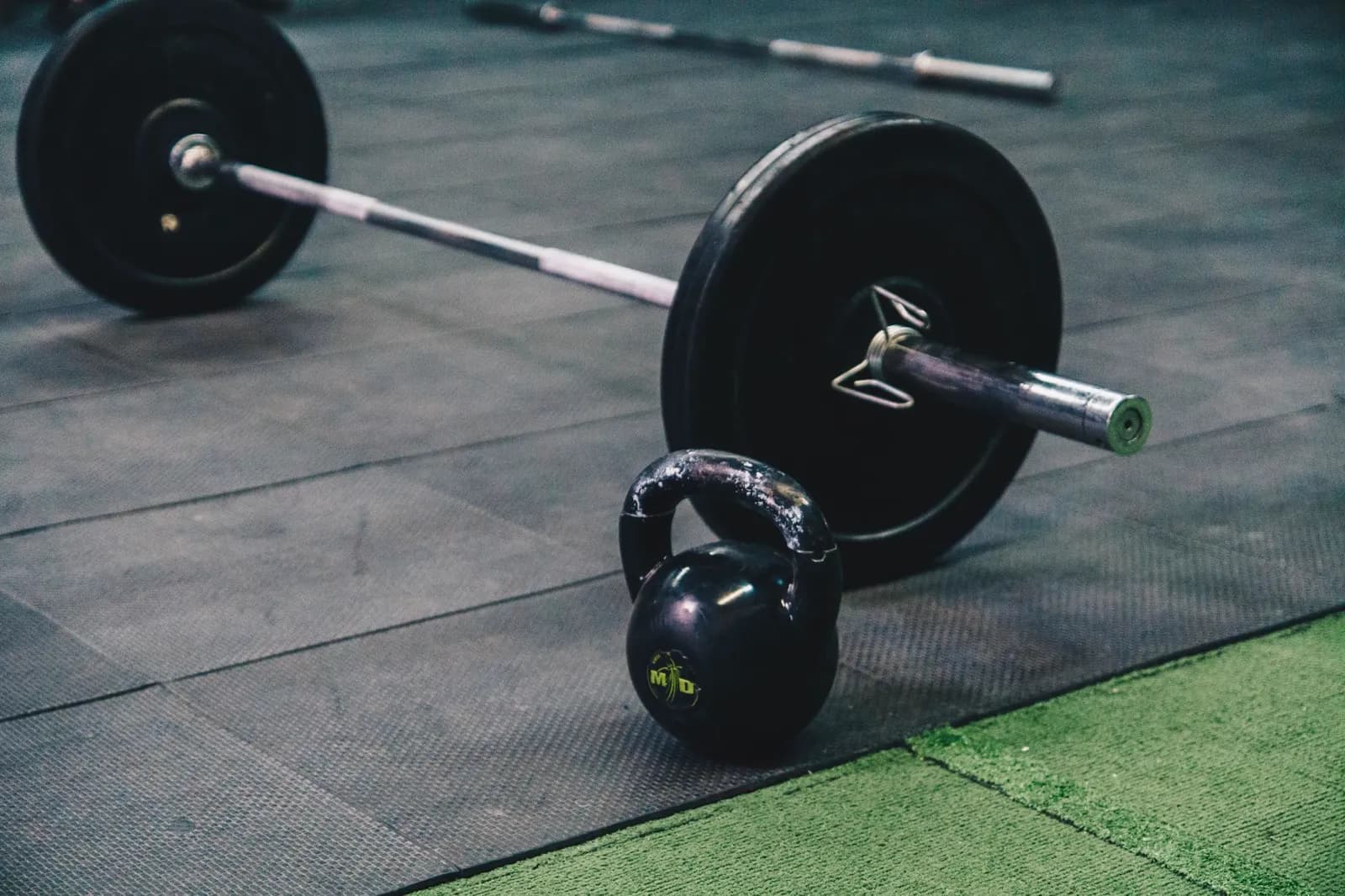A loaded barbell and a kettlebell resting on the rubber matting of a free-weights floor, beside a strip of green artificial turf.