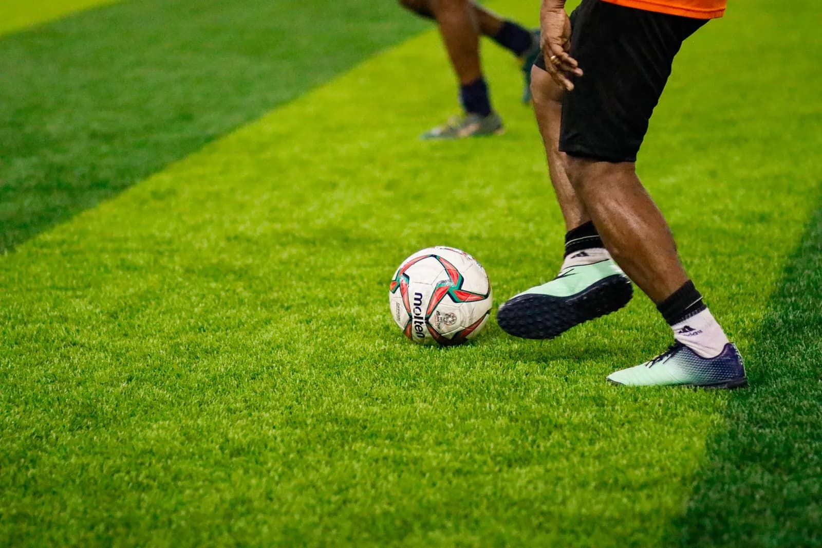 A player's legs and a match ball on brightly lit artificial turf, with a second player running in the background.