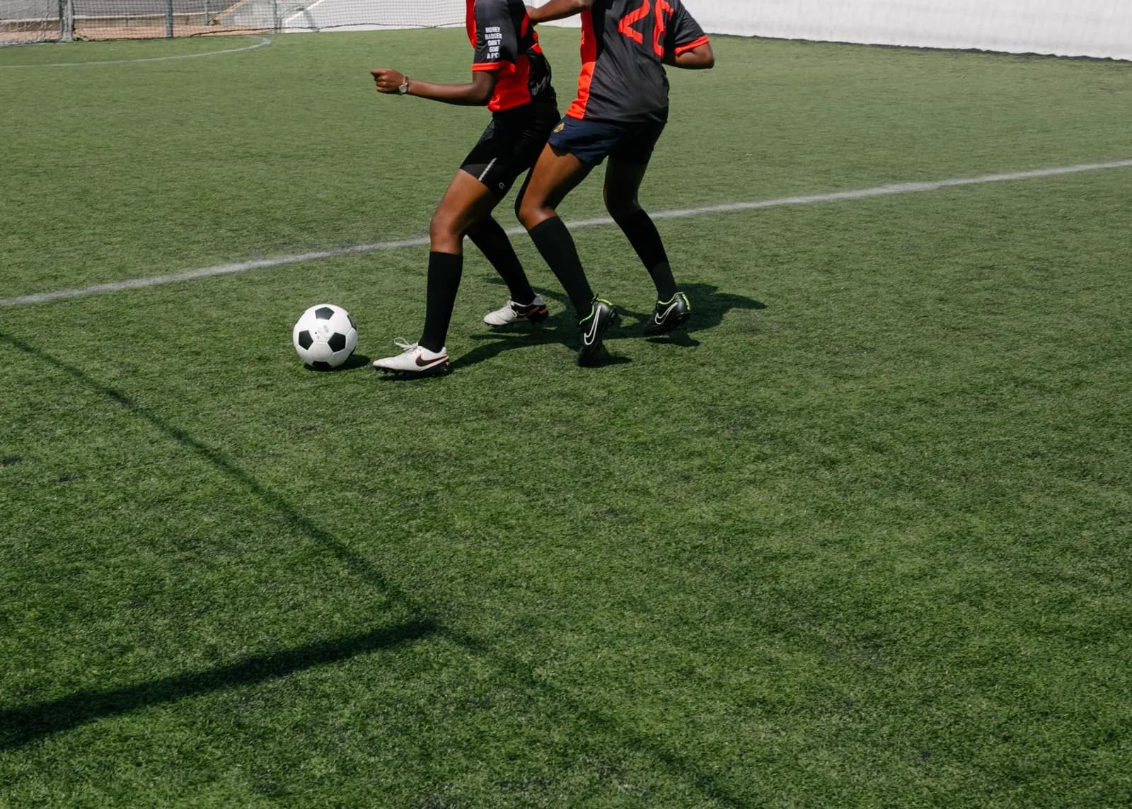 Two players in kit contesting the ball on marked artificial turf, cropped at the shoulders, with perimeter fencing behind them.