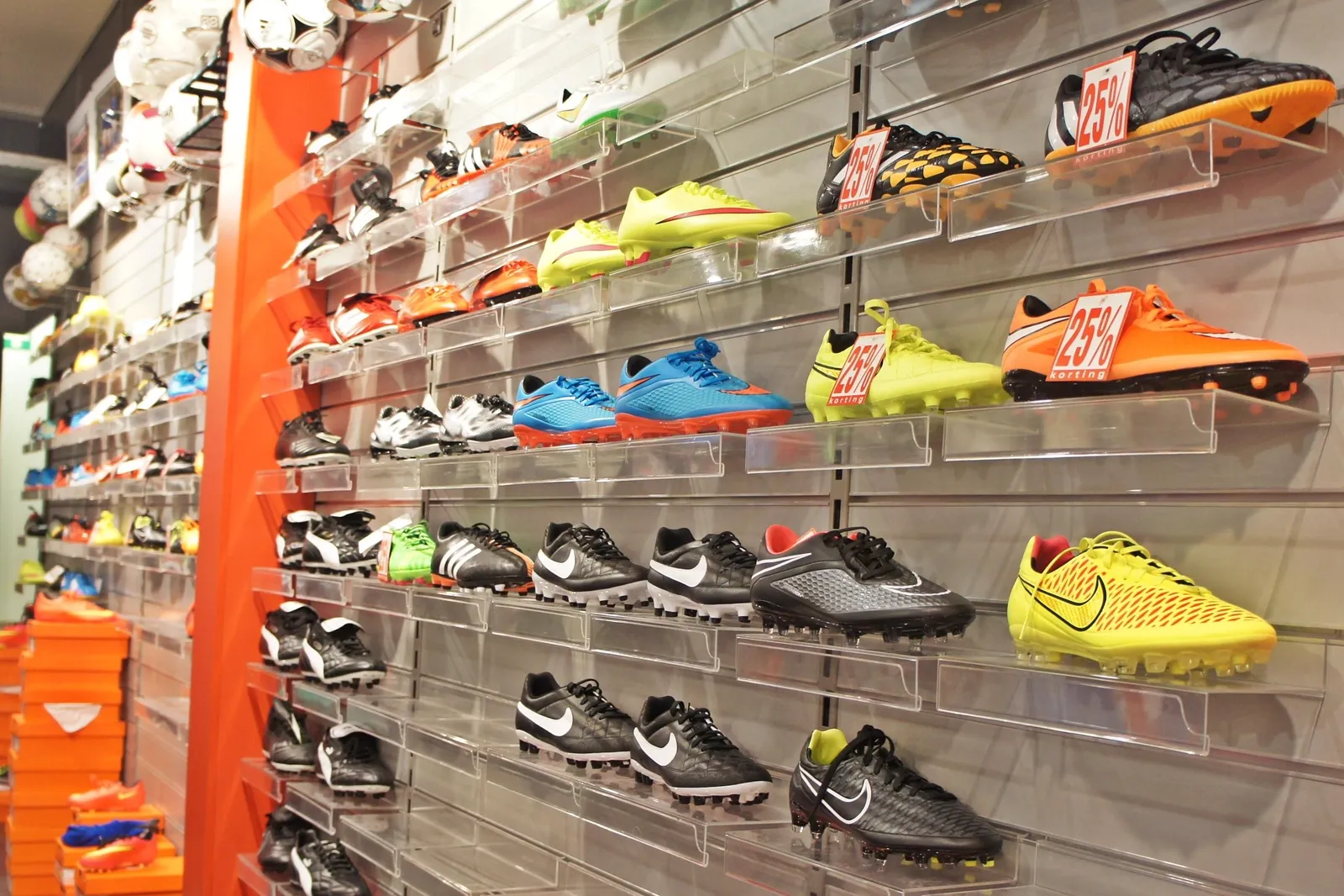 A retail wall of football boots on clear acrylic shelves, with footballs racked above and stacked shoeboxes to one side.